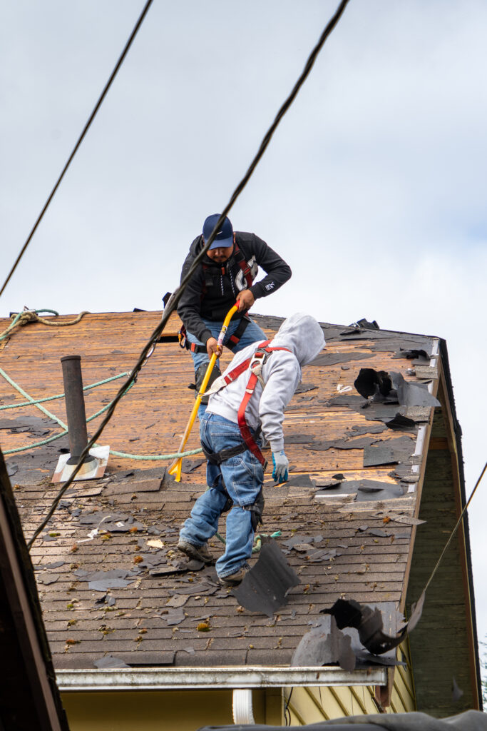 Two workers repairing a damaged roof with shovels and safety harnesses.