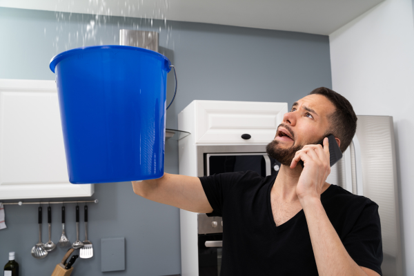 A worried man holding a blue bucket to catch water from a ceiling leak while on the phone.