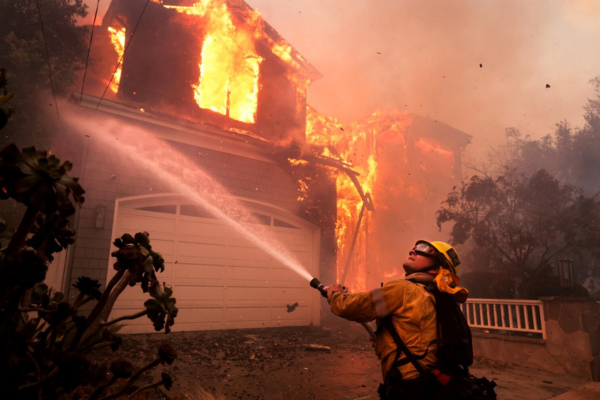 A firefighter sprays water on a burning home, surrounded by thick smoke and flames.